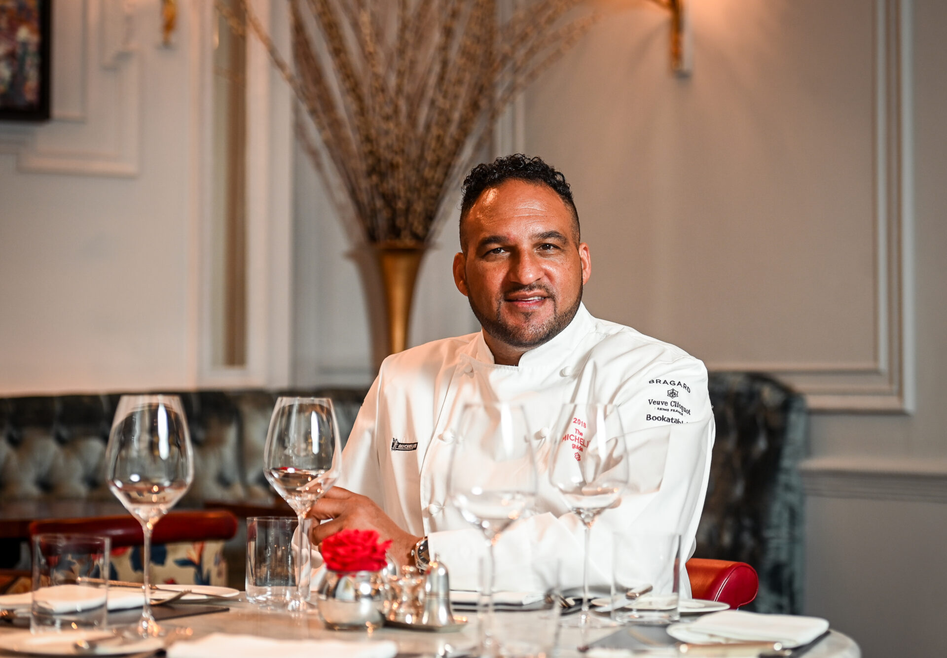 A chef in a white jacket sits at a table set with empty wine glasses, plates, and a red rose centerpiece in the elegant dining room of The Stafford London, surrounded by soft lighting and decorative wall accents.