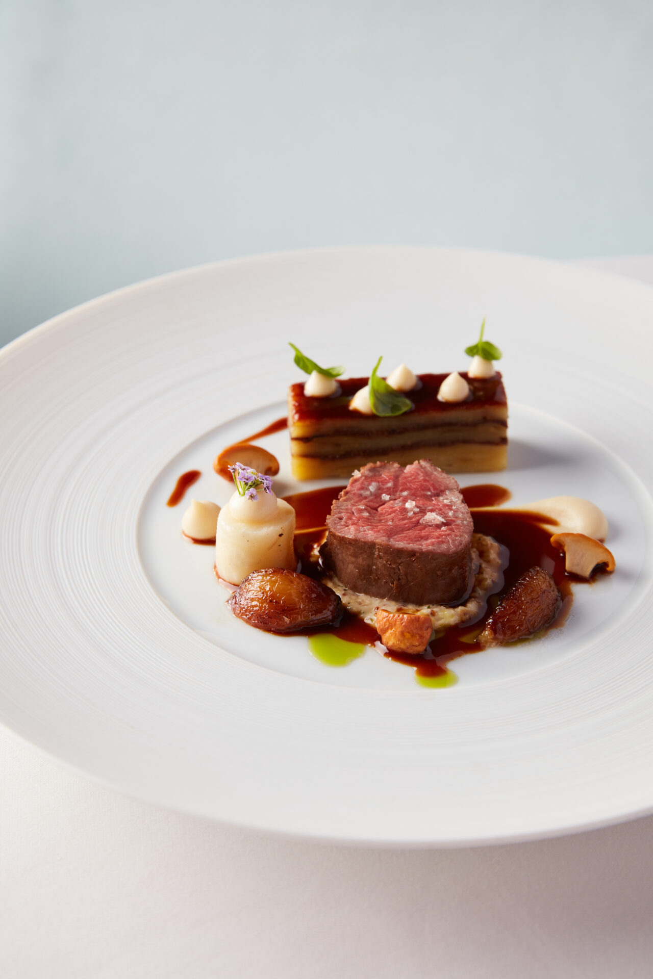 A gourmet dish from The Game Bird featuring a round cut of medium-rare beef, layered potato, dollops of sauce, and garnishes, all artfully arranged on a white plate with a delicate drizzle of brown sauce.