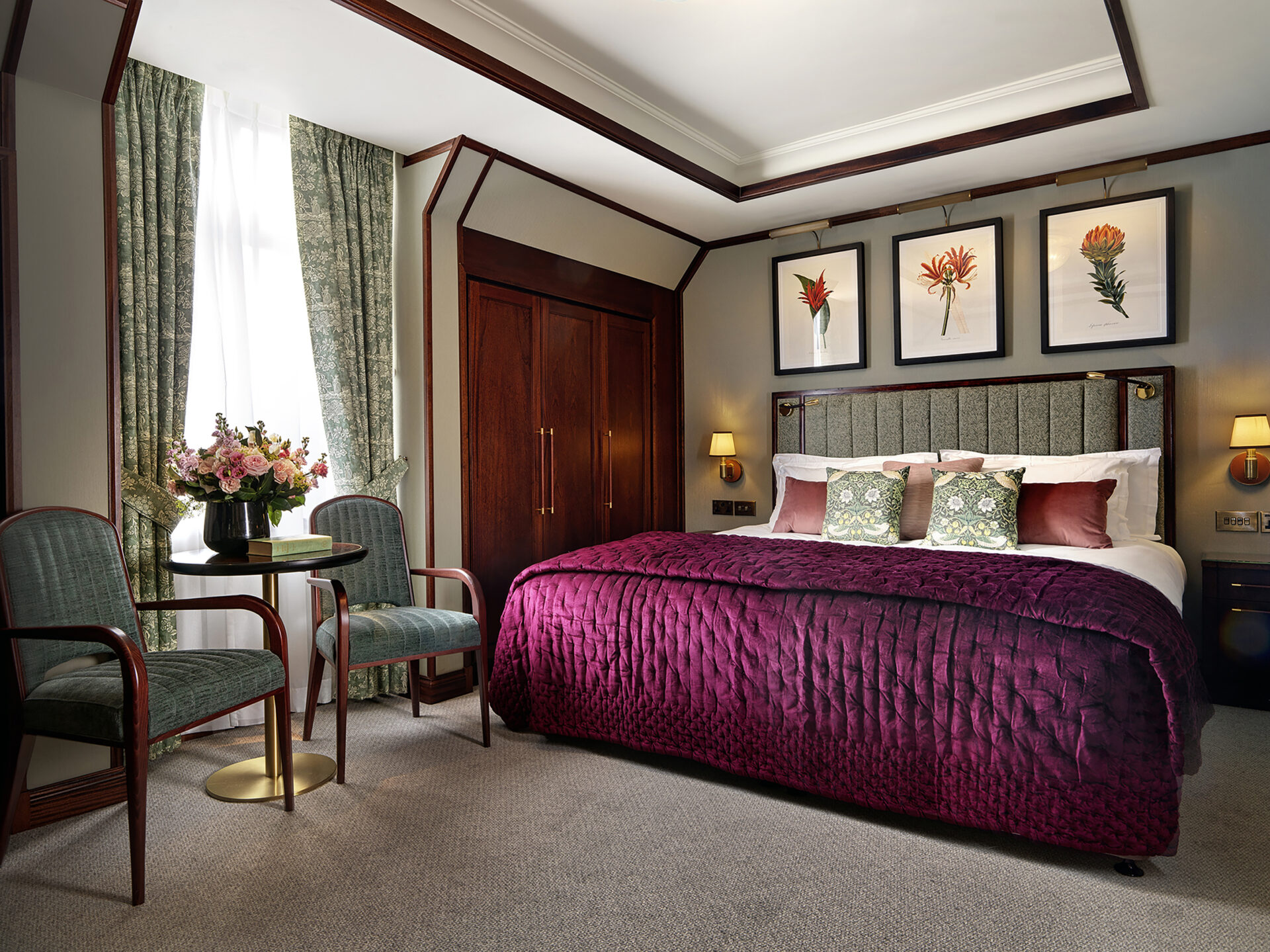 A stylish hotel room with a large bed covered in a deep purple quilt, decorative pillows, two green chairs with a small round table, floral artwork above the bed, and natural light from a window with green drapes.