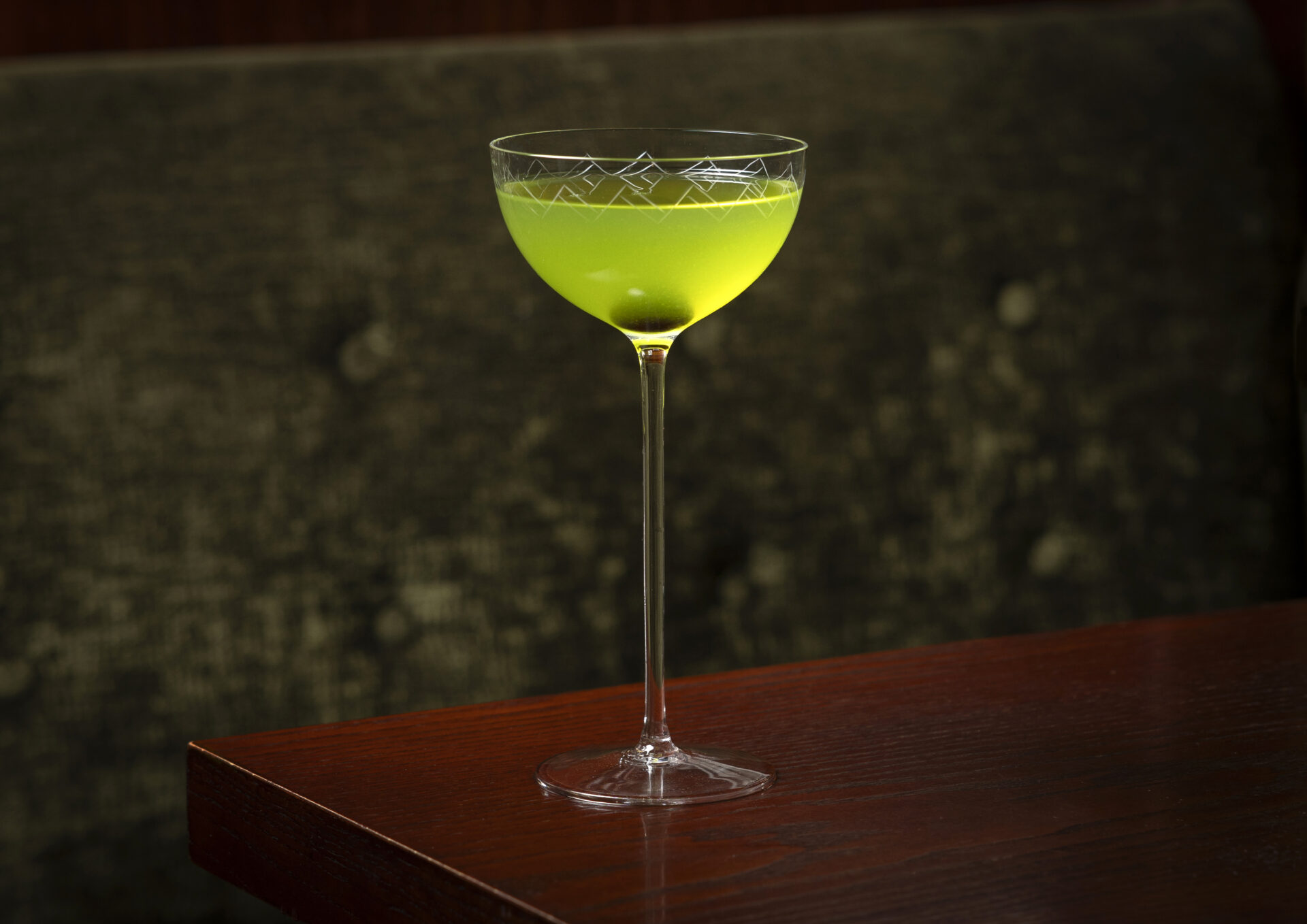 A coupe glass filled with a bright green cocktail from The American Bar sits on a dark wooden table with a textured, dark-colored background.