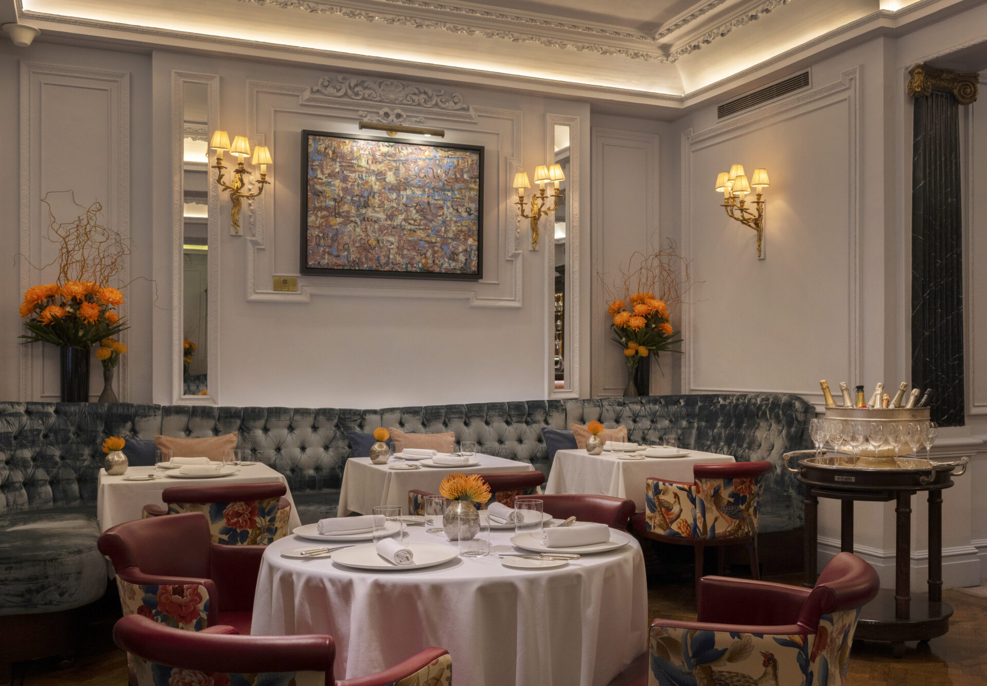 Elegant restaurant with white tablecloths, red chairs, and blue tufted banquette seating. At Michael Caines at The Stafford, tables are set with plates and glasses, orange flowers brighten the space, and art adorns paneled walls.