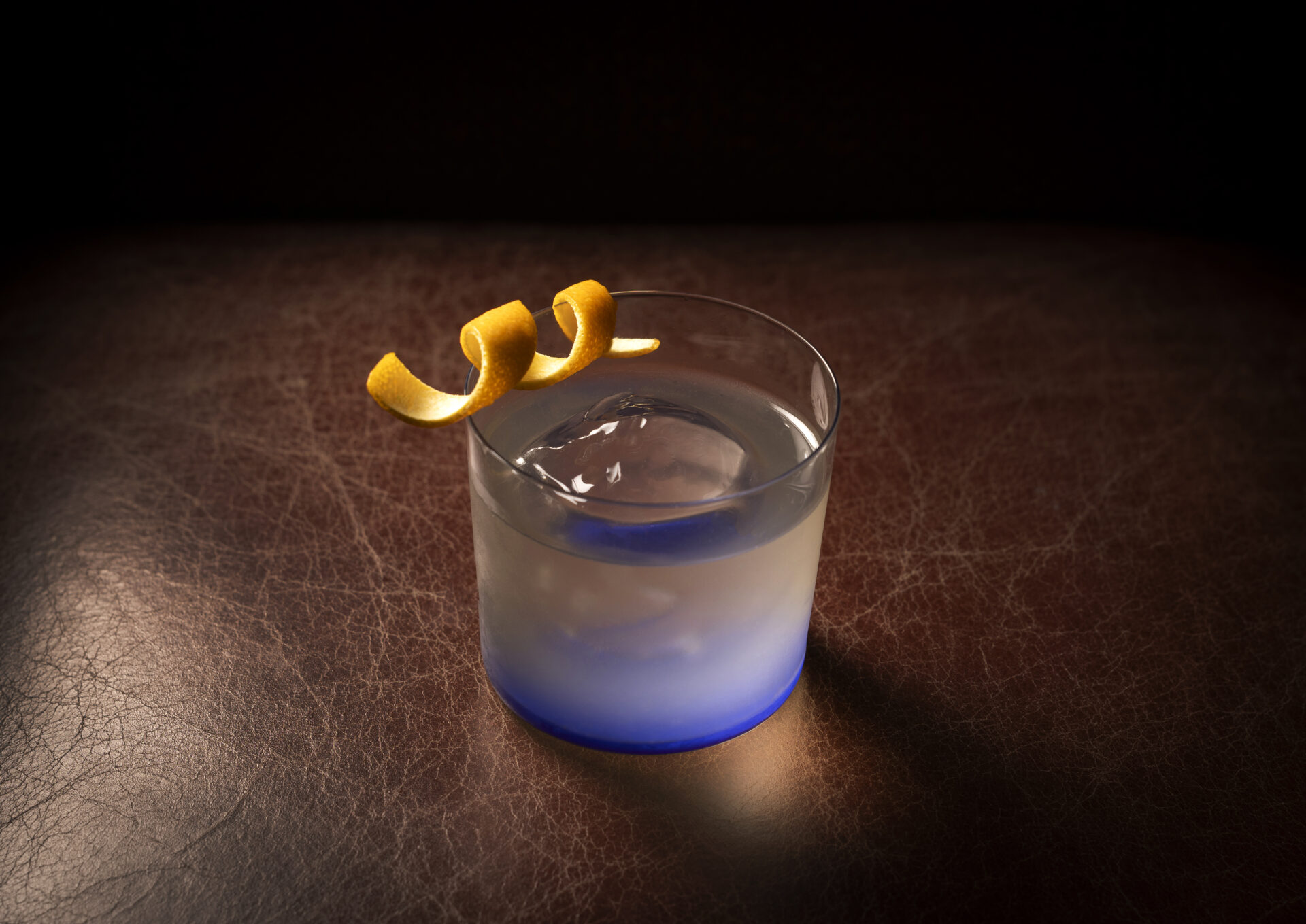 A clear cocktail in a low glass with a large ice sphere, garnished with a curled orange peel, sits on a dark brown leather surface under dramatic lighting—an elegant creation inspired by The American Bar.