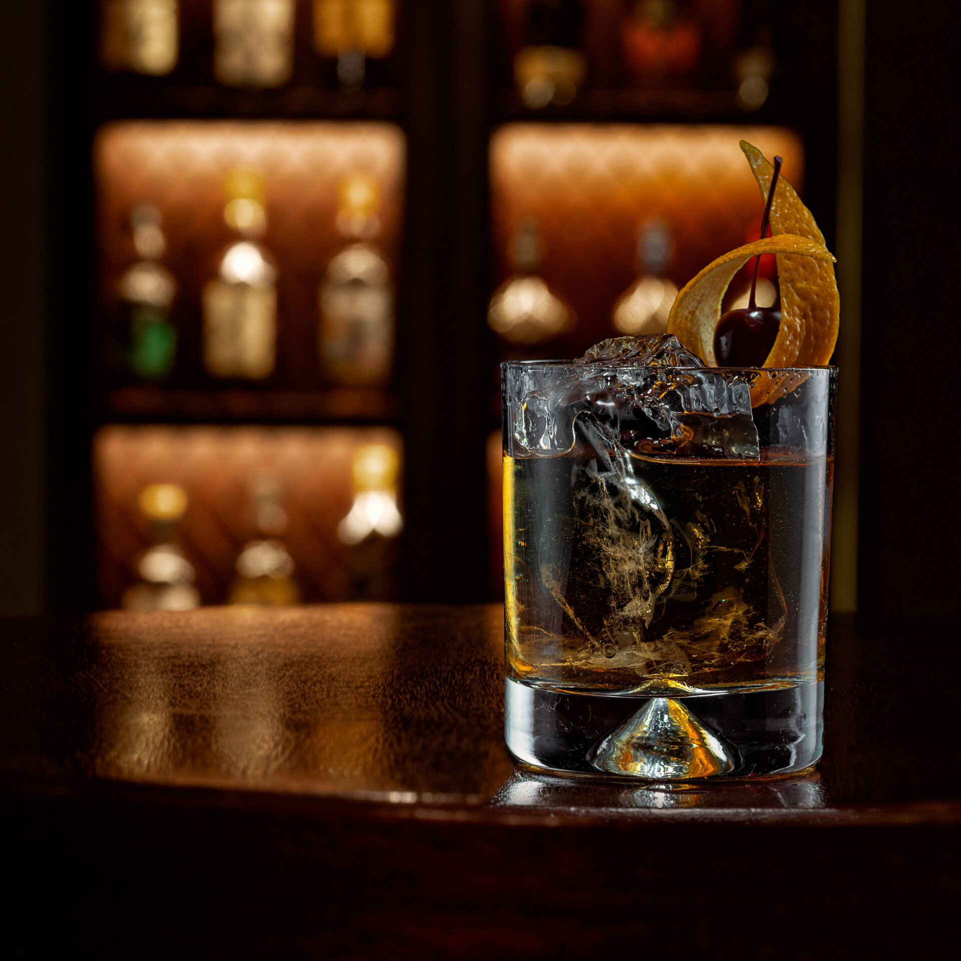 A glass of whiskey with ice, garnished with an orange peel and cherry, rests on a polished bar counter at The American Bar, with shelves of liquor bottles softly lit in the blurred background.