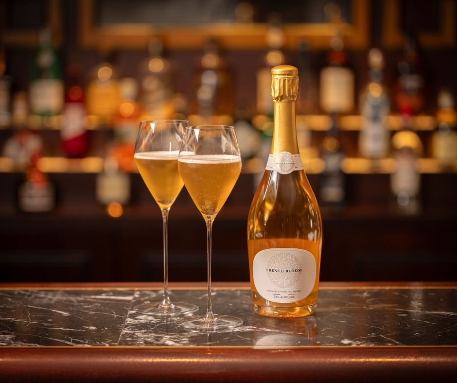 A bottle of sparkling wine and two filled wine glasses sit on a dark marble bar, with blurred bottles of liquor visible on shelves in the background.
