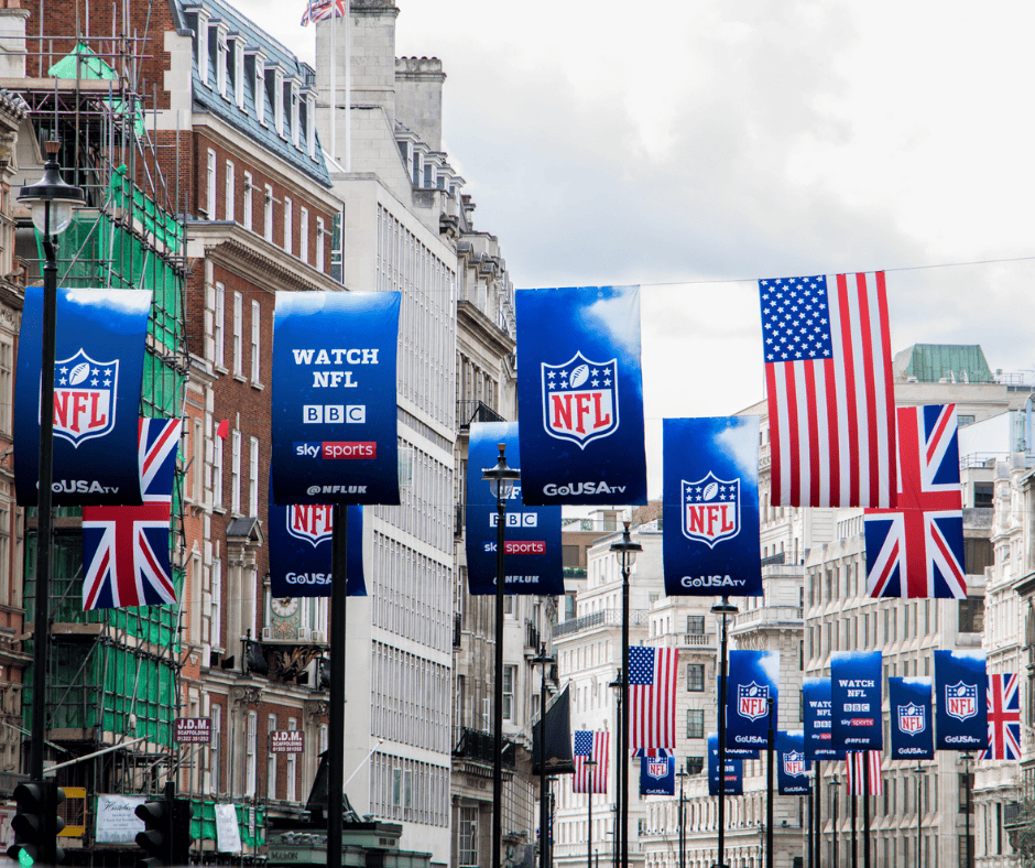 A city street lined with NFL banners and British and American flags. Classic buildings flank both sides, with signs for The American Bar and to watch NFL on BBC, Sky Sports, and Go USA TV. The sky is mostly cloudy.